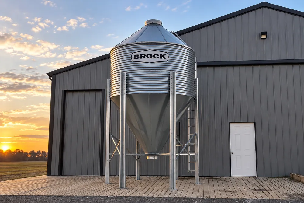 Brock hopper-bottom grain bin beside a farm building at sunset.