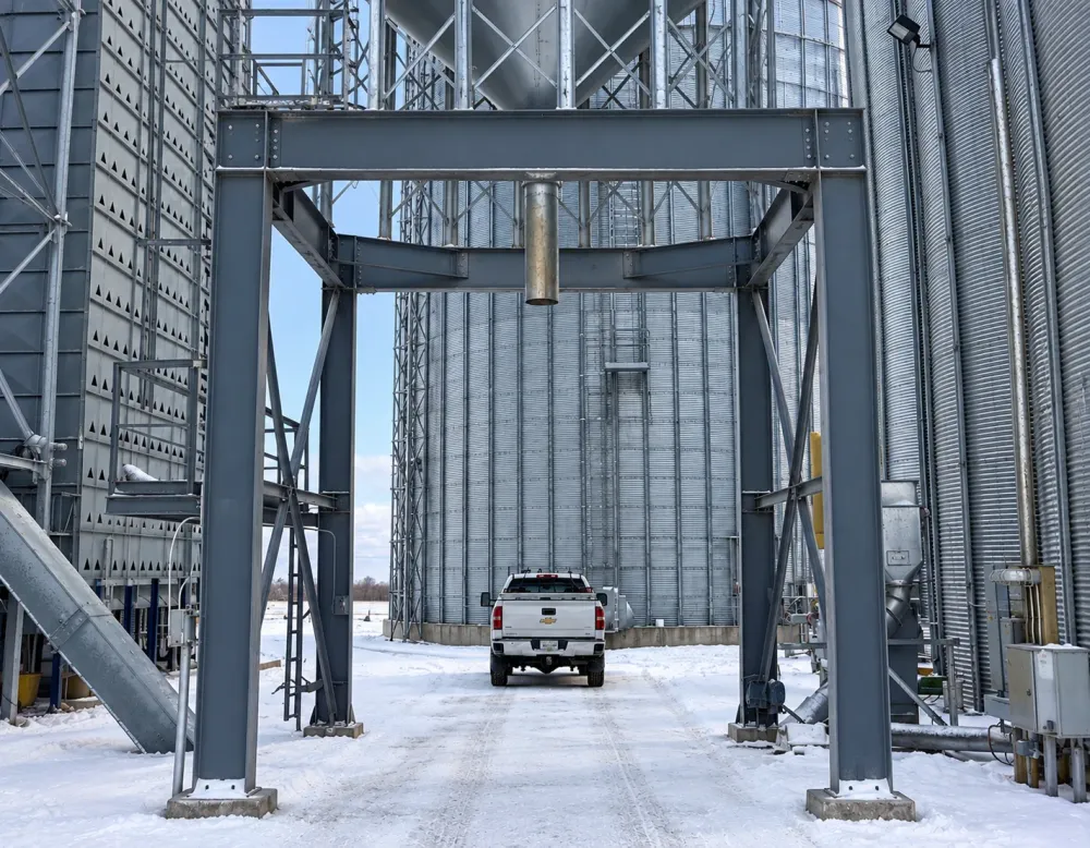 Steel support frame and overhead hopper between grain storage bins, with a pickup truck beneath.